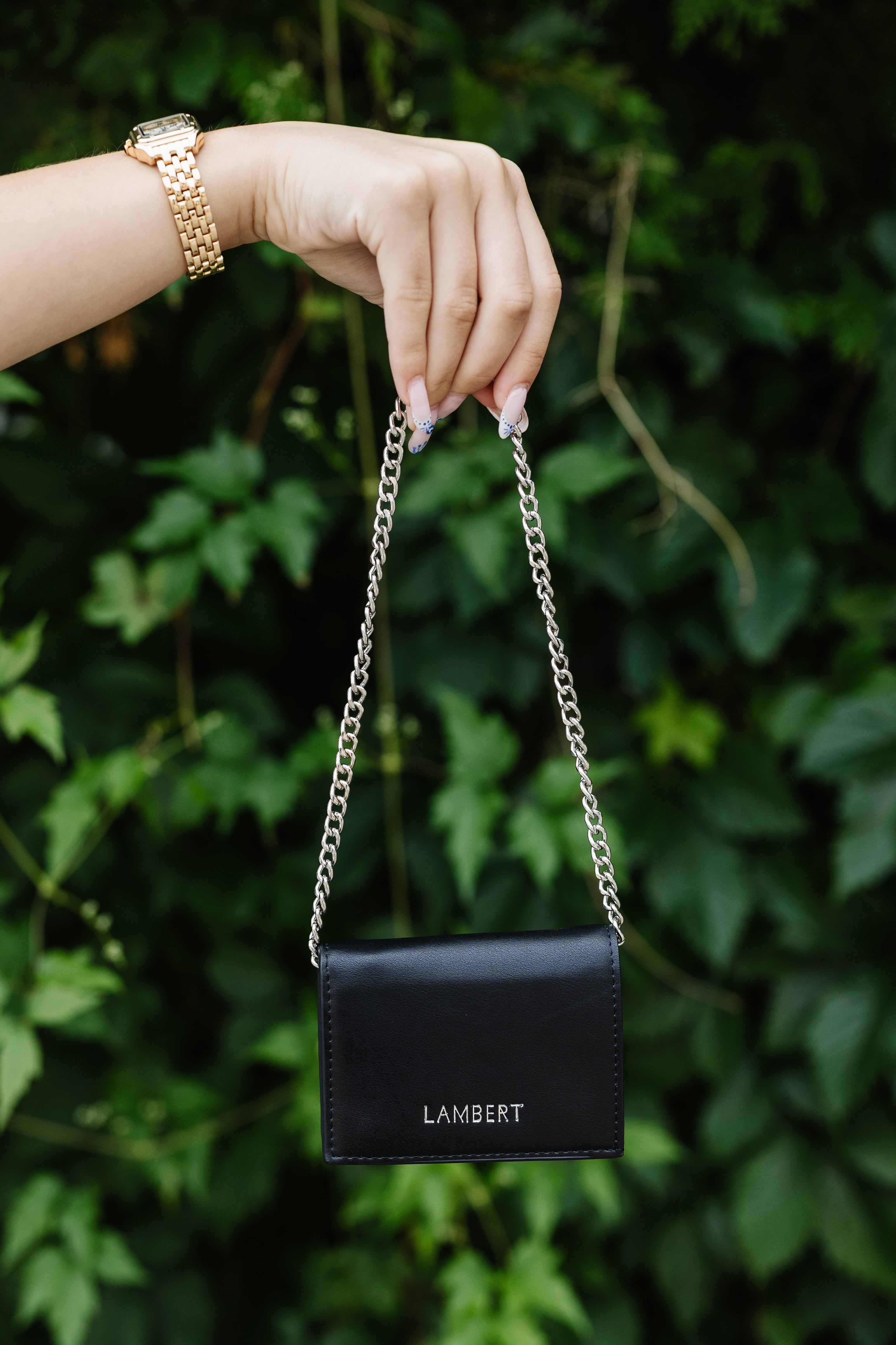 Black handbag with chain strap held by a hand against a green leafy background
