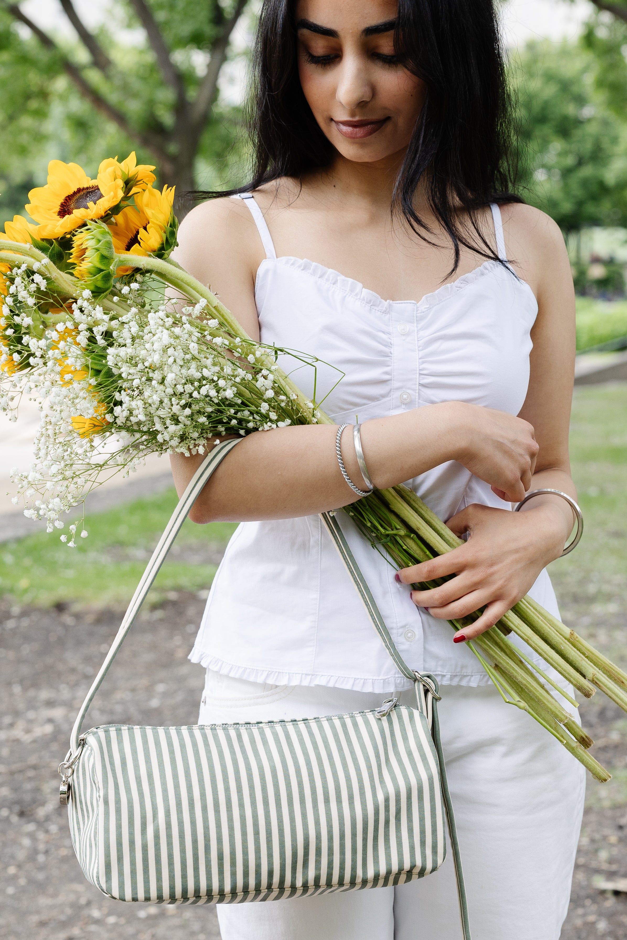 A woman in a white sleeveless top and white pants holds a striped shoulder bag and a bouquet of sunflowers and baby's breath outdoors.