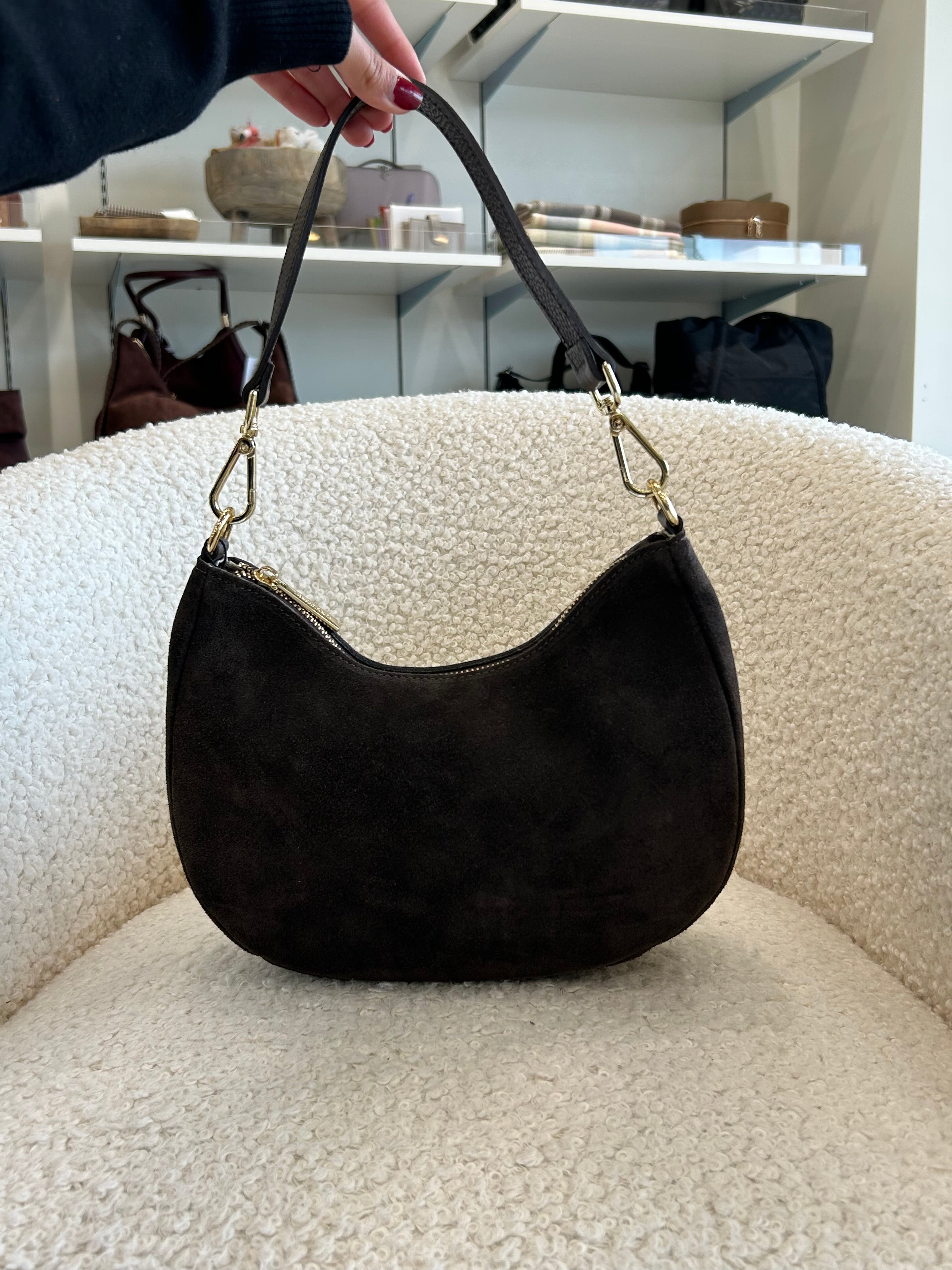 A dark brown suede crescent-shaped shoulder bag with gold-tone hardware, held by a hand over a cream-colored boucle armchair in a boutique setting.