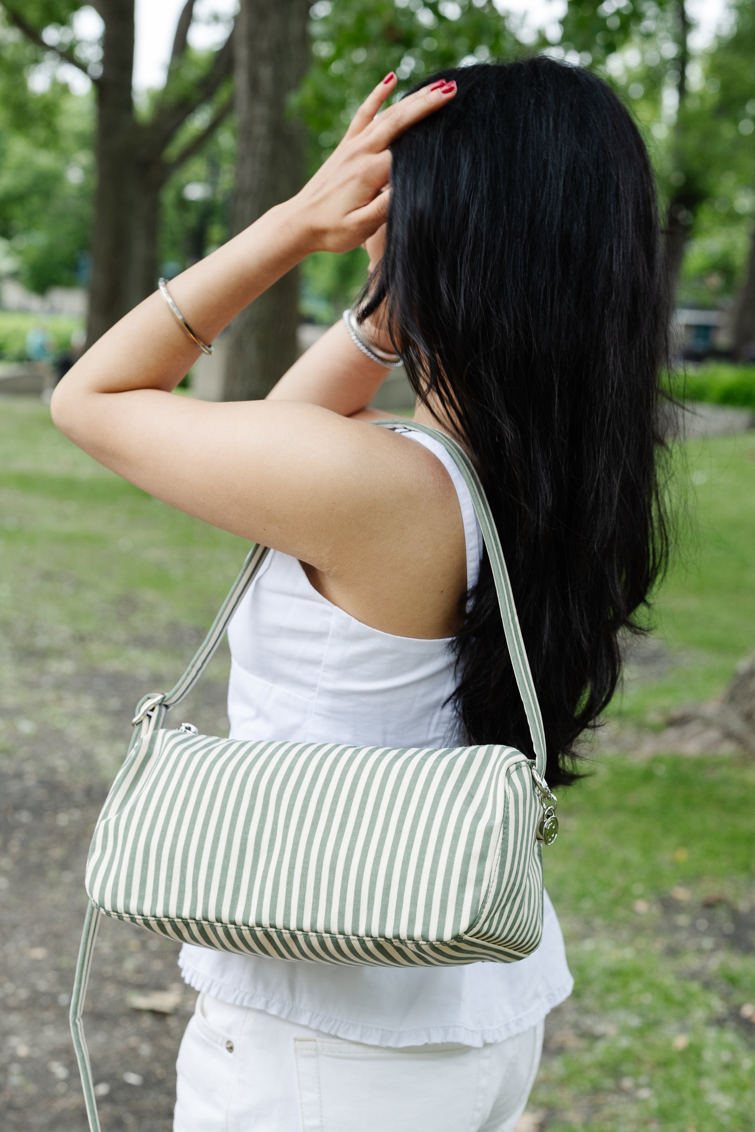 A woman with long black hair is shown from behind in an outdoor park setting, wearing a white sleeveless top and white pants while carrying a green and white vertically striped shoulder bag.